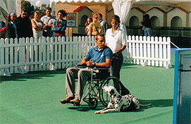 Enrique Solís, director de LealCan, en ua exhibición de perros de asistencia. Él es llevado en silla de ruedas por una mujer y al lado de la silla, un perro dálmata está tumbado y lo mira, pendiente