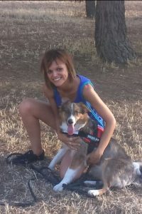 Educadora canina apoyando su pierna izquierda en la hierba y abrazando a su perra Anya que está a su izquierda con las dos manos