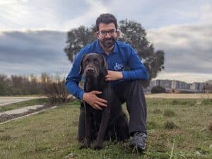 Enrique Fernández apoyando la pierna derecha en la hierba, abrazando a su perro Broccoli que se encuentra sentado delante suyo, ambos mirando a la cámara